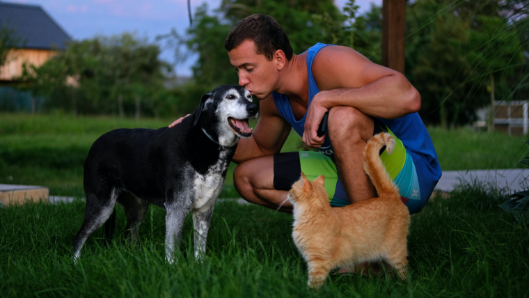 A man embraces a pet dog while a ginger cat looks on.